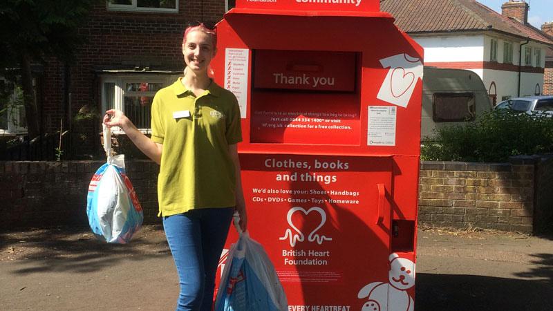 A Student Community Warden stands in front of a British Heart Foundation red donation bin with two bags of donations